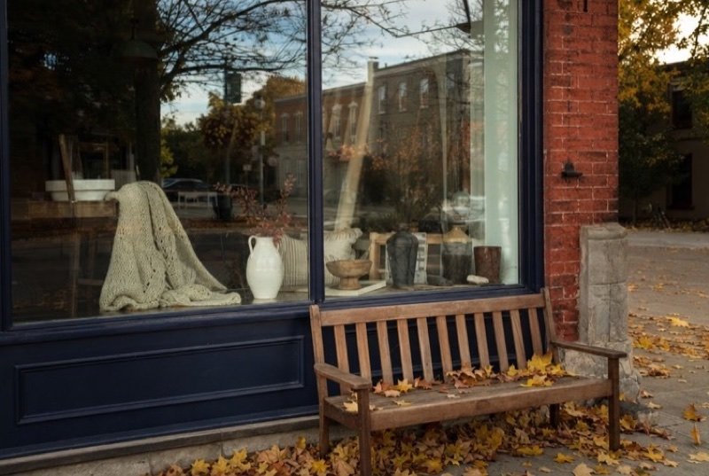 The window of an independent boutique in a red-brick building, a wooden bench outside and maple leaves on the pavement