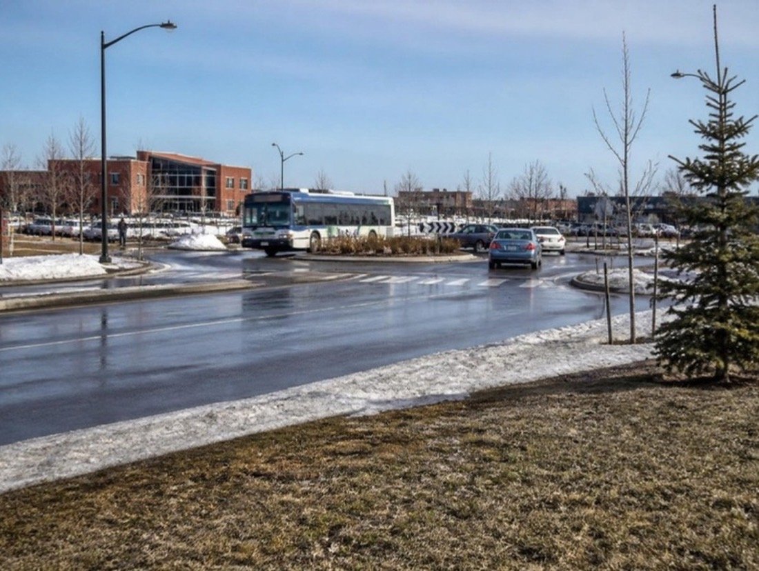 A busy four-lane commercial road on a bright March morning, a landscaped roundabout ahead