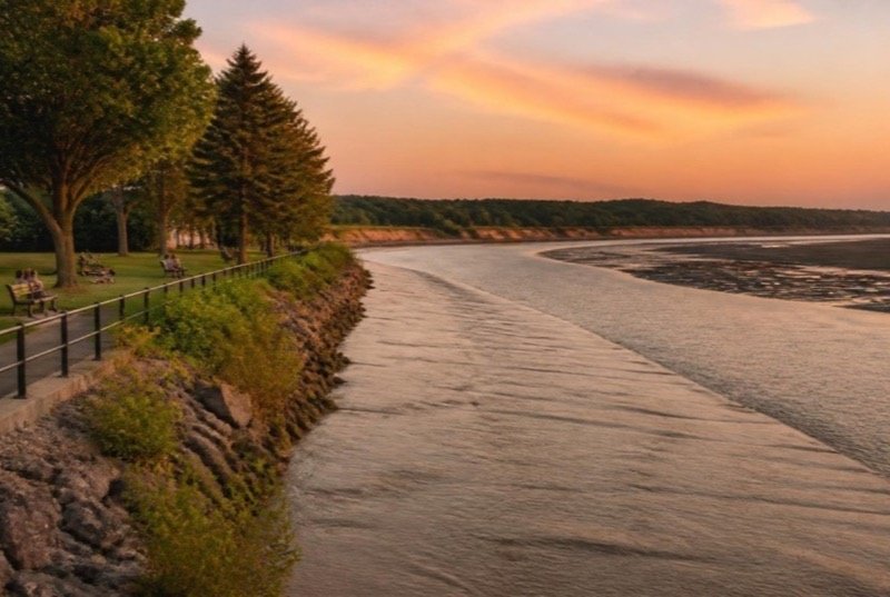 A wide silt-brown tidal river at golden hour, a low wave front moving upriver over pale mudflats beside a grassy park with a paved trail