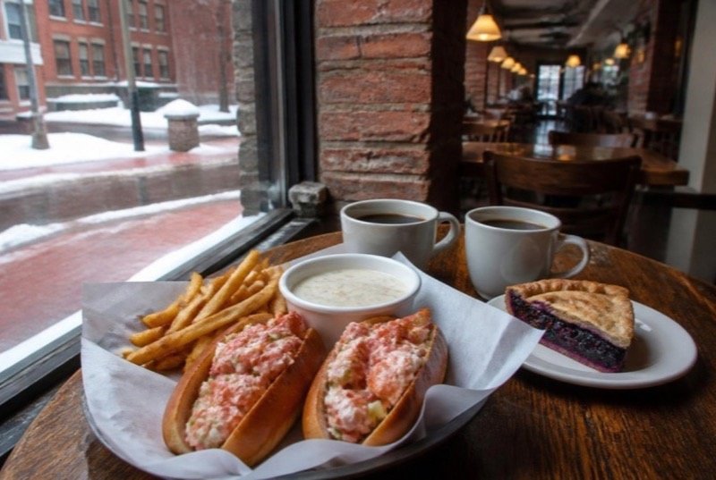 A paper-lined tray of two lobster rolls with fries, a bowl of chowder and coffees on a wooden table by a tall window onto a snowy brick street