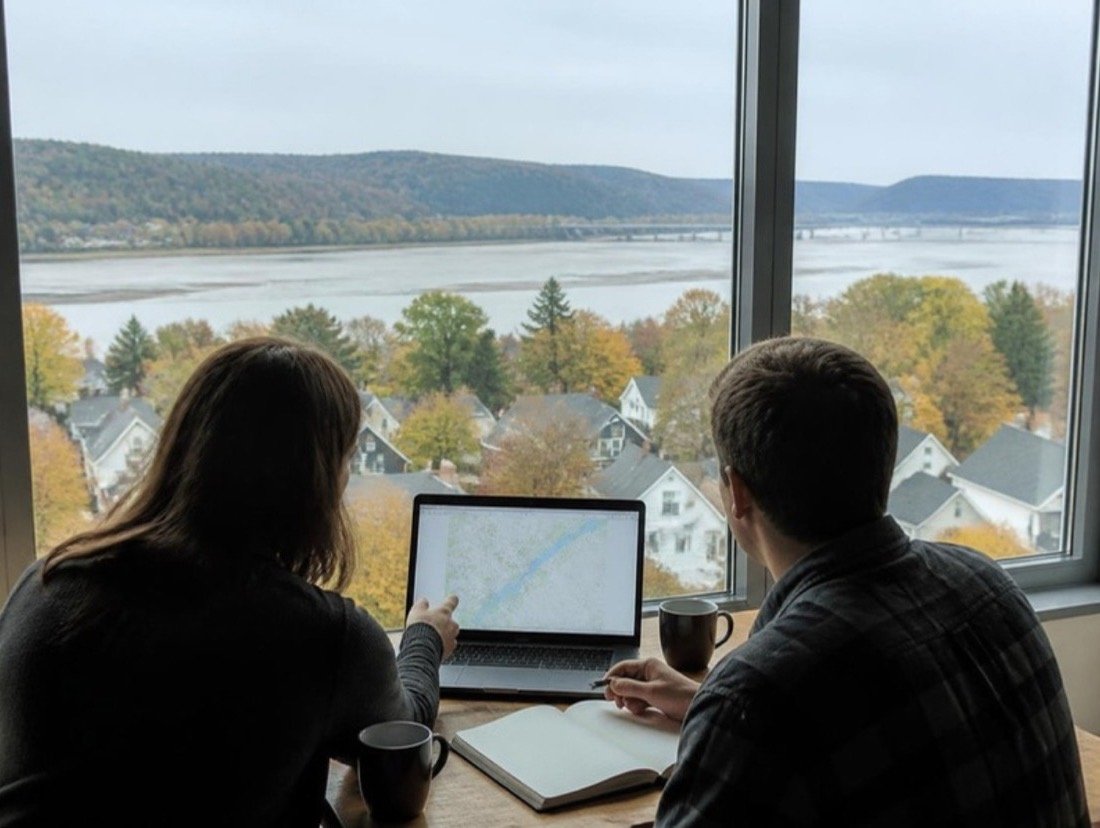 Seen from behind, two people at a wooden table with a laptop map and an open notebook, a river valley beyond the window